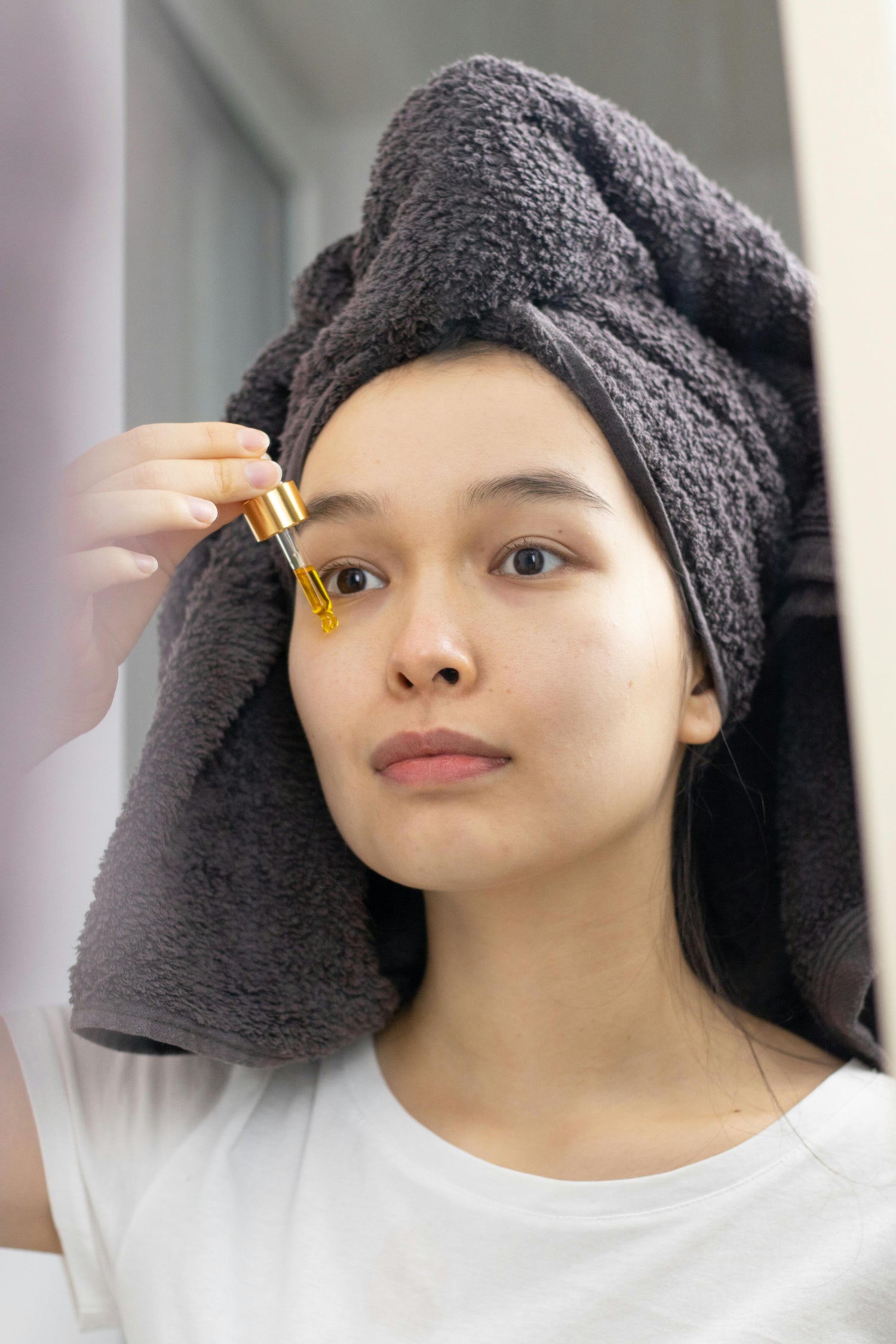 Women applying a dropper of skincare product to their face with a towel on their head.